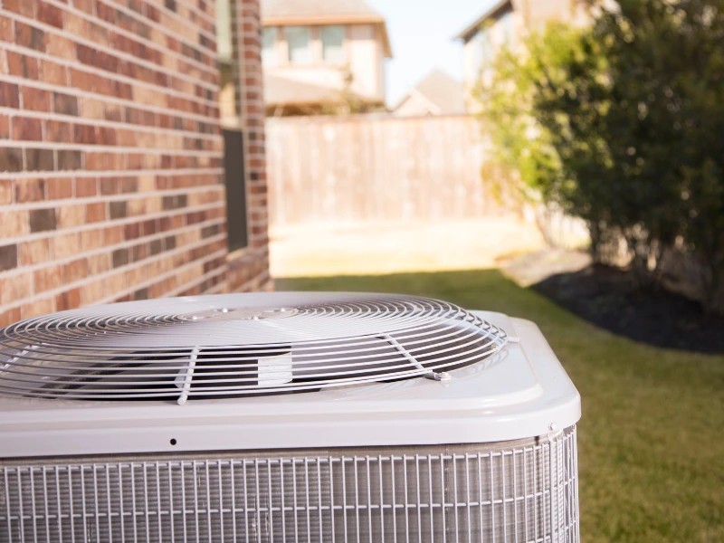 Outdoor air conditioner sits behind house shaded from direct sunlight.